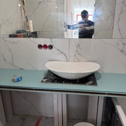 STROY BUD.group ANDRII PYLYPENKO - Montaż umywalki nablatowej w łazience z marmurowymi płytkami; widoczne narzędzia, poziomica, odbicie pracownika w lustrze.
