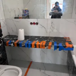 Półka z czarnego marmuru w łazience w trakcie montażu, zabezpieczona pomarańczową i niebieską taśmą, białe płytki imitujące marmur na ścianach, odbicie osoby robiącej zdjęcie w lustrze.