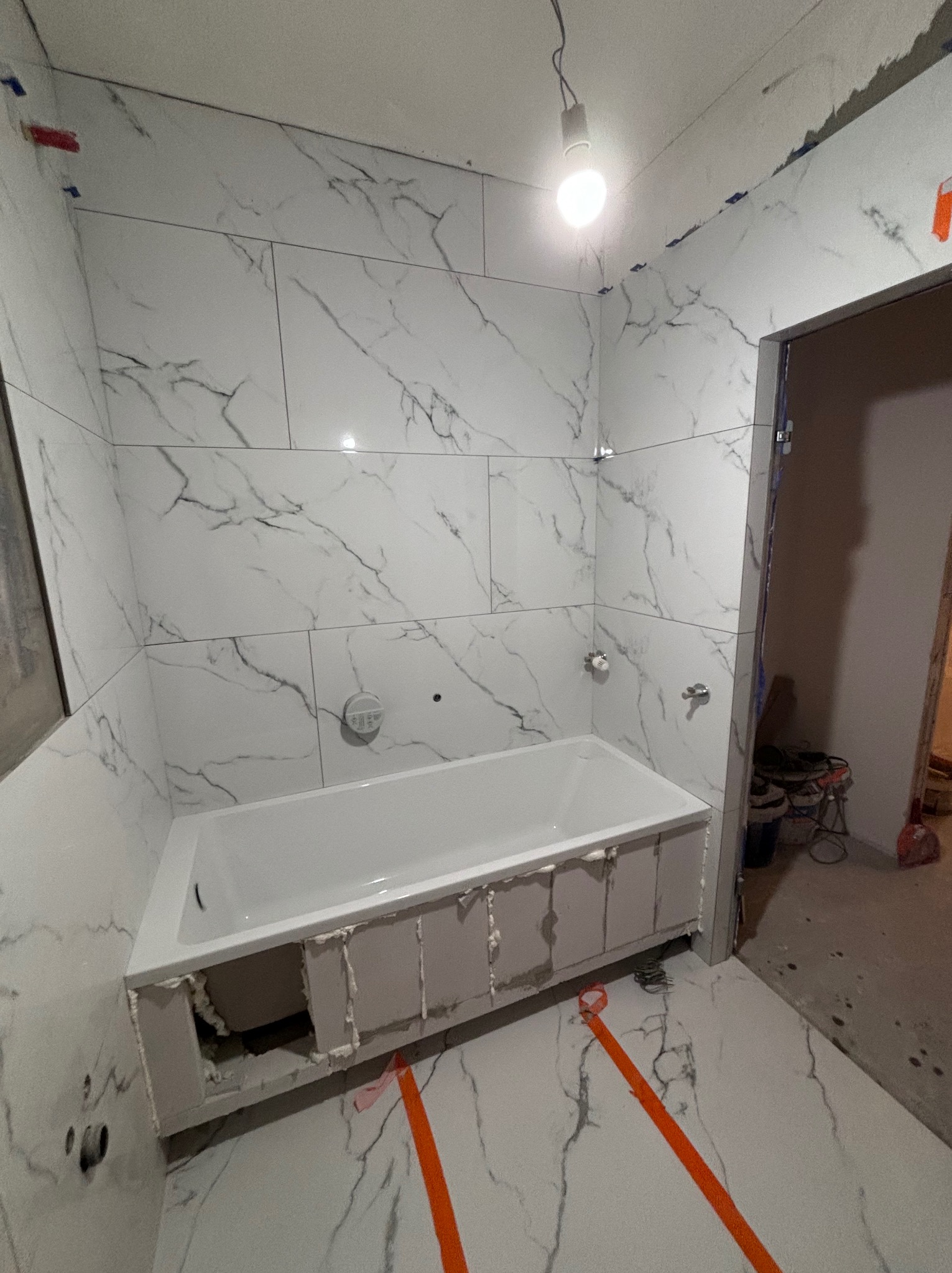 Biała wanna obudowana płytami gipsowymi, częściowo wykończona, otoczona marmurowymi płytkami na ścianach i podłodze, z widocznymi śladami prac budowlanych i pomarańczową taśmą zabezpieczającą.