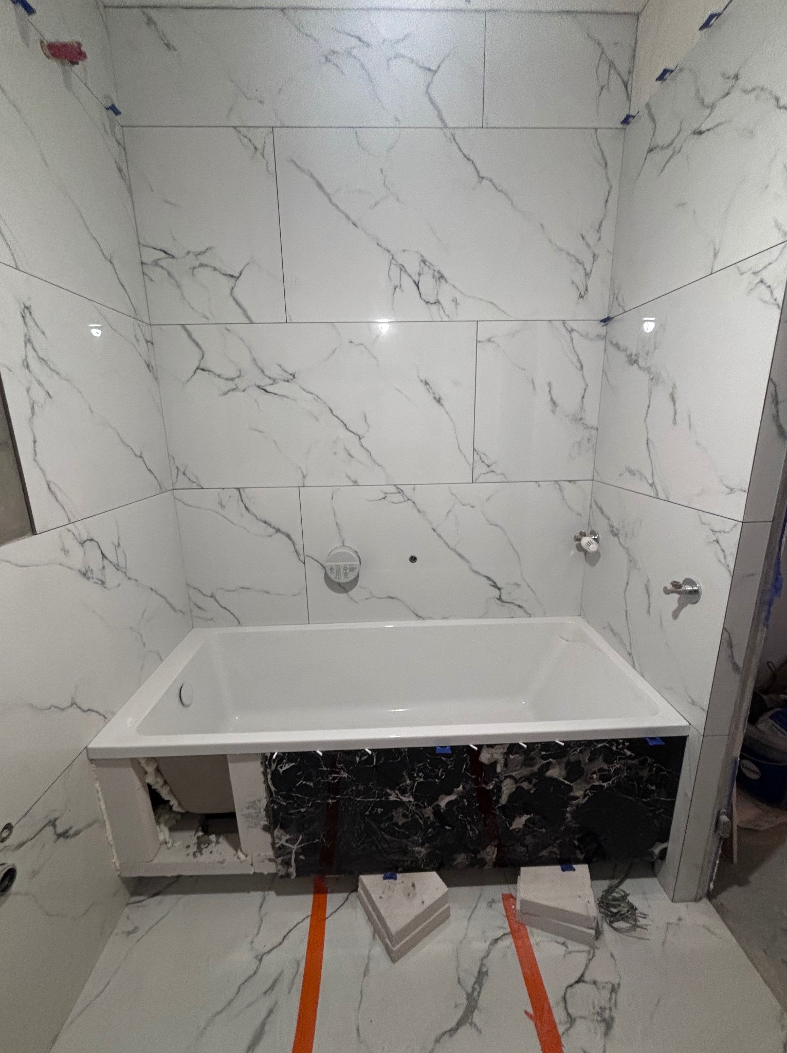 Montaż wanny z czarnym marmurem na obudowie, białe płytki imitujące marmur na ścianach i podłodze, widoczne elementy instalacji wodnej, etap prac wykończeniowych.