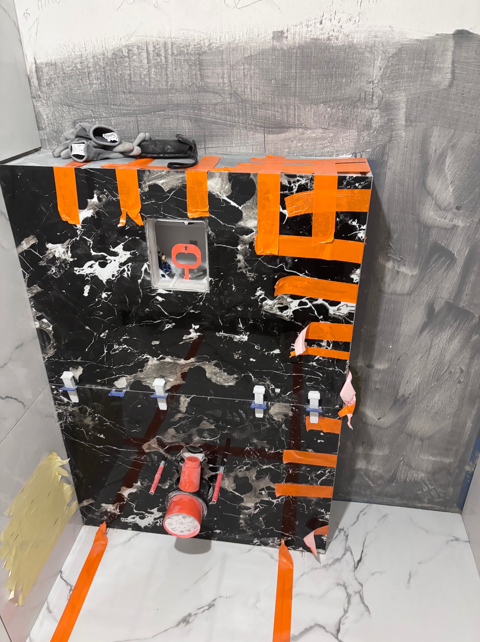 Montaż stelaża podtynkowego WC z obudową imitującą czarny marmur, zabezpieczonego pomarańczową taśmą malarską, widoczne białe płytki podłogowe z szarym wzorem marmuru, szare ściany w trakcie...