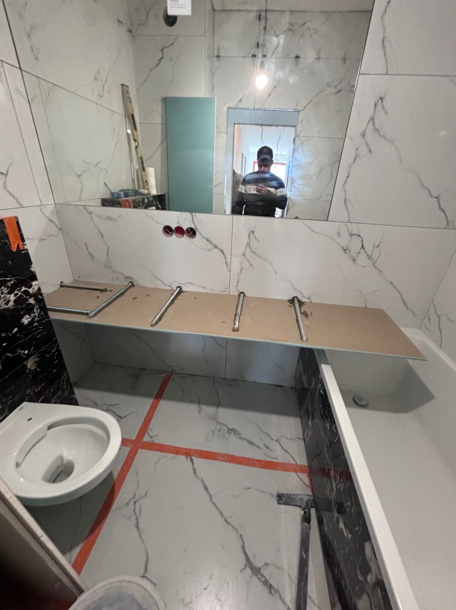 Łazienka w trakcie remontu: białe płytki imitujące marmur, zamontowany stelaż pod umywalkę, częściowo widoczna toaleta, pomarańczowe linie wyznaczające układ płytek na podłodze, odbicie osoby...