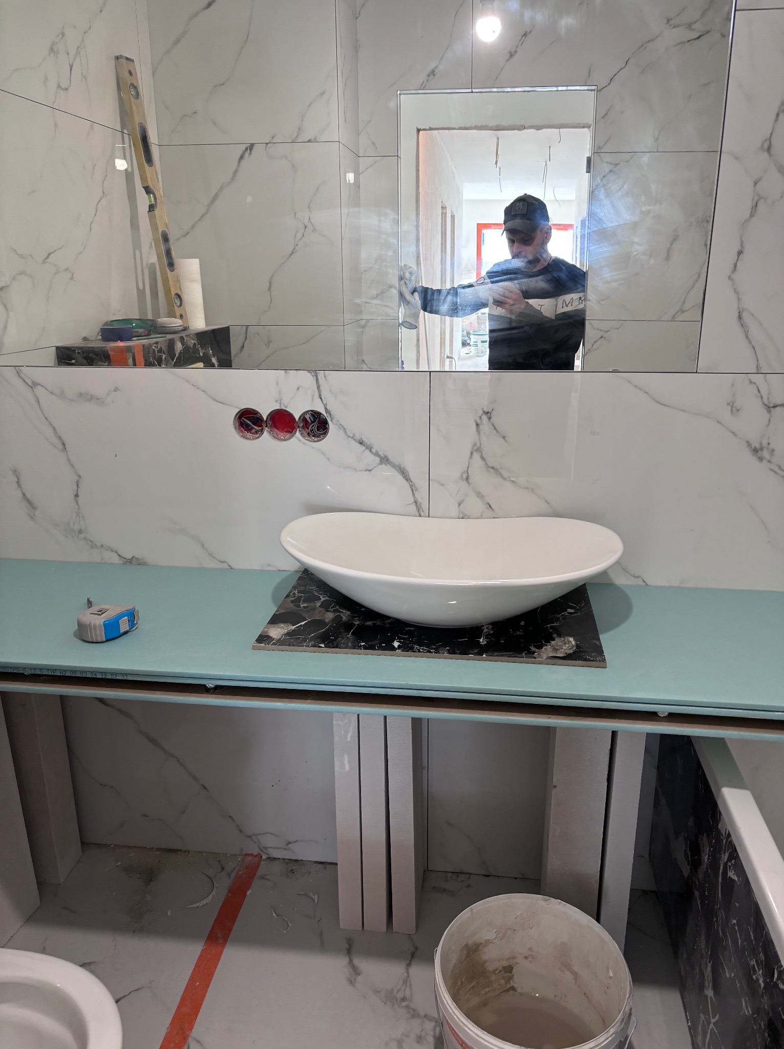 Montaż umywalki nablatowej w łazience z marmurowymi płytkami; widoczne narzędzia, poziomica, odbicie pracownika w lustrze.