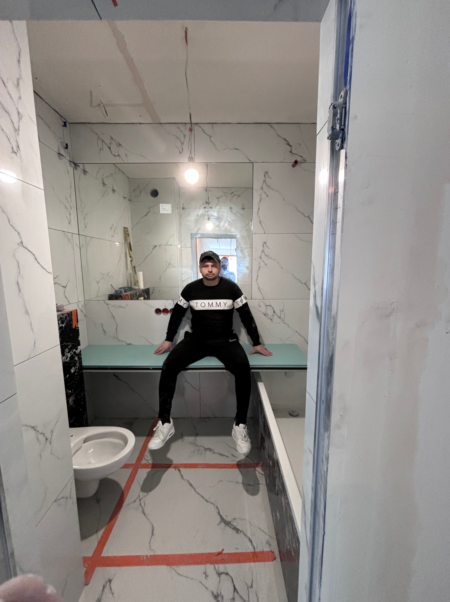 Wnętrze łazienki w trakcie prac wykończeniowych, z marmurowymi płytkami na ścianach i podłodze, widoczna toaleta, obudowa wanny, oraz mężczyzna siedzący na obudowie wanny, wyznaczona strefa taśmą...