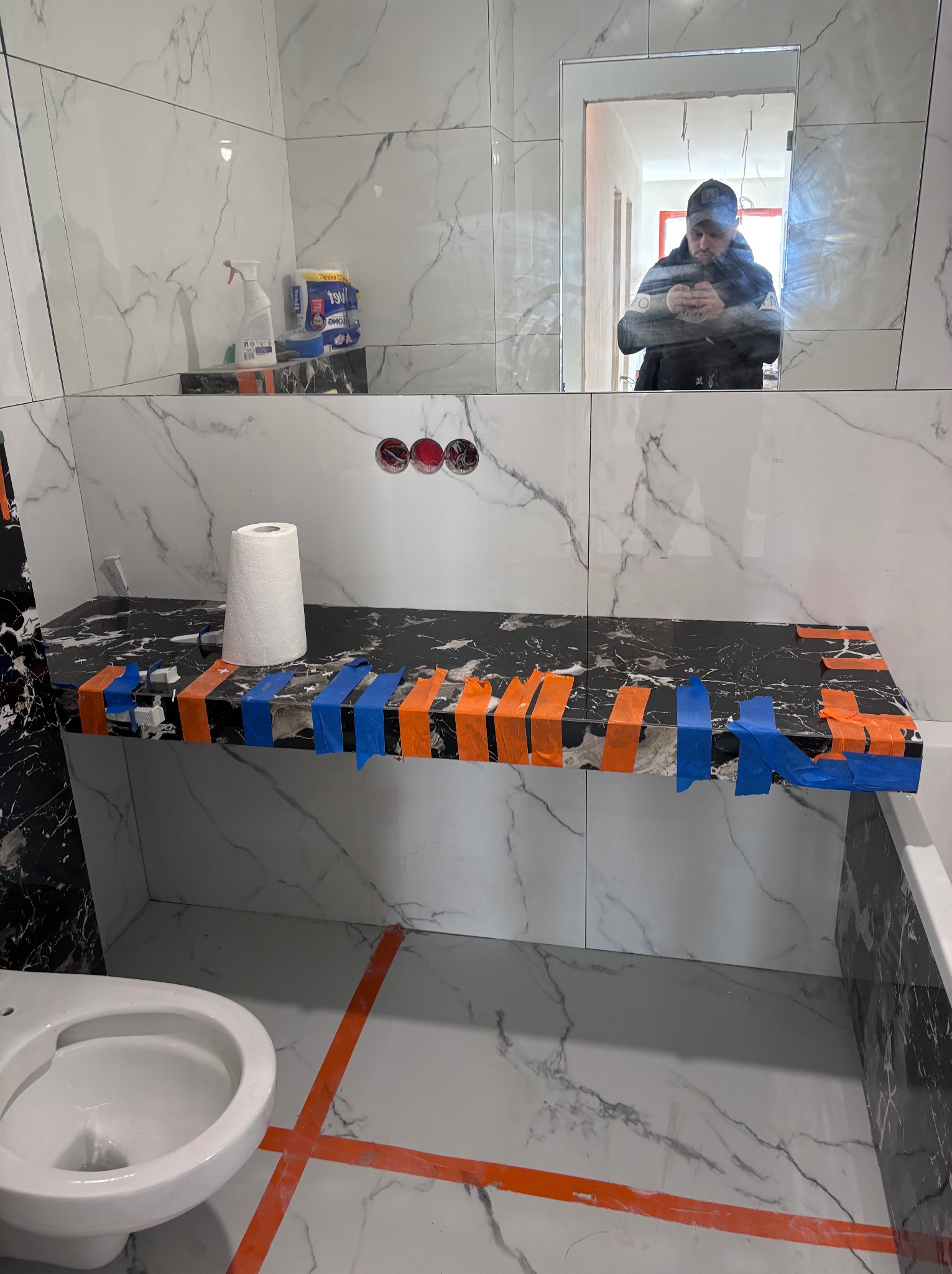 Półka z czarnego marmuru w łazience w trakcie montażu, zabezpieczona pomarańczową i niebieską taśmą, białe płytki imitujące marmur na ścianach, odbicie osoby robiącej zdjęcie w lustrze.