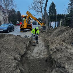 WODKAN GNIECH SPÓŁKA Z OGRANICZONĄ ODPOWIEDZIALNOŚCIĄ - Pracownik w zielonej kamizelce zagęszcza ziemię w wykopie przy użyciu ubijarki spalinowej, w tle koparka JCB i ulica z samochodami.