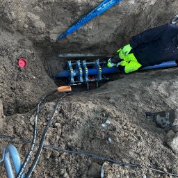 WODKAN GNIECH SPÓŁKA Z OGRANICZONĄ ODPOWIEDZIALNOŚCIĄ - Pracownik w odblaskowej kurtce łączy rury wodociągowe w wykopie za pomocą specjalistycznego zaciskarki hydraulicznej, widoczne kable hydrauliczne i taśma ostrzegawcza.