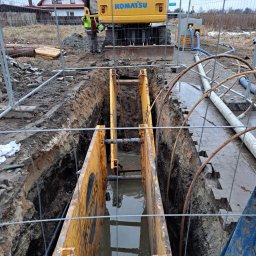 WODKAN GNIECH SPÓŁKA Z OGRANICZONĄ ODPOWIEDZIALNOŚCIĄ - Głęboki wykop zabezpieczony żółtymi szalunkami, wypełniony wodą, z pracującą koparką Komatsu w tle i widocznym pracownikiem w odblaskowej kamizelce, teren budowy ogrodzony siatką.