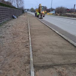 WODKAN GNIECH SPÓŁKA Z OGRANICZONĄ ODPOWIEDZIALNOŚCIĄ - Przygotowanie podłoża pod chodnik z kostki brukowej, wyrównany piasek i obrzeża z kostki betonowej, w tle koparka i pracownicy.