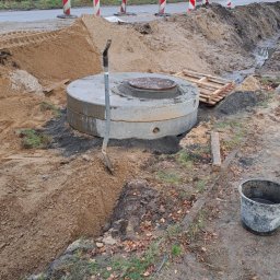 WODKAN GNIECH SPÓŁKA Z OGRANICZONĄ ODPOWIEDZIALNOŚCIĄ - Wkopany betonowy krąg z włazem kanalizacyjnym, obok łopata wbita w ziemię, widoczne zabezpieczenia drogowe i logo firmy WOD-KAN Gniech.