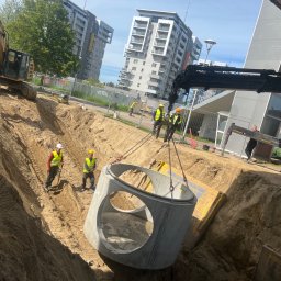 WODKAN GNIECH SPÓŁKA Z OGRANICZONĄ ODPOWIEDZIALNOŚCIĄ - Montaż betonowego kręgu z otworami na rury w wykopie, z użyciem dźwigu, pracownicy w kamizelkach odblaskowych, na tle budynki mieszkalne.