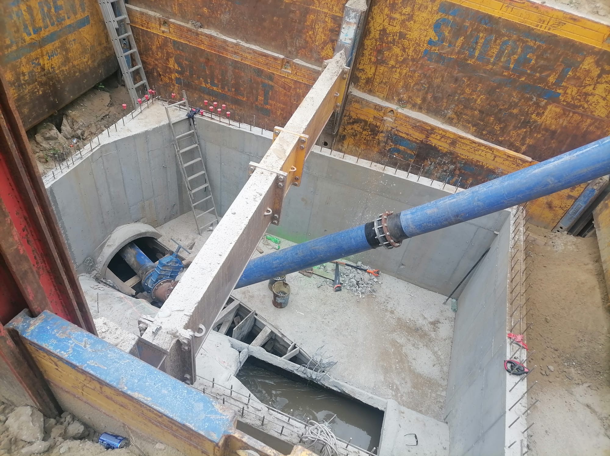 Głęboki wykop z betonowymi ścianami, zabezpieczony żółtymi szalunkami, z widoczną rurą doprowadzającą i drabinkami zejściowymi, fragment kałuży na dnie wykopu.