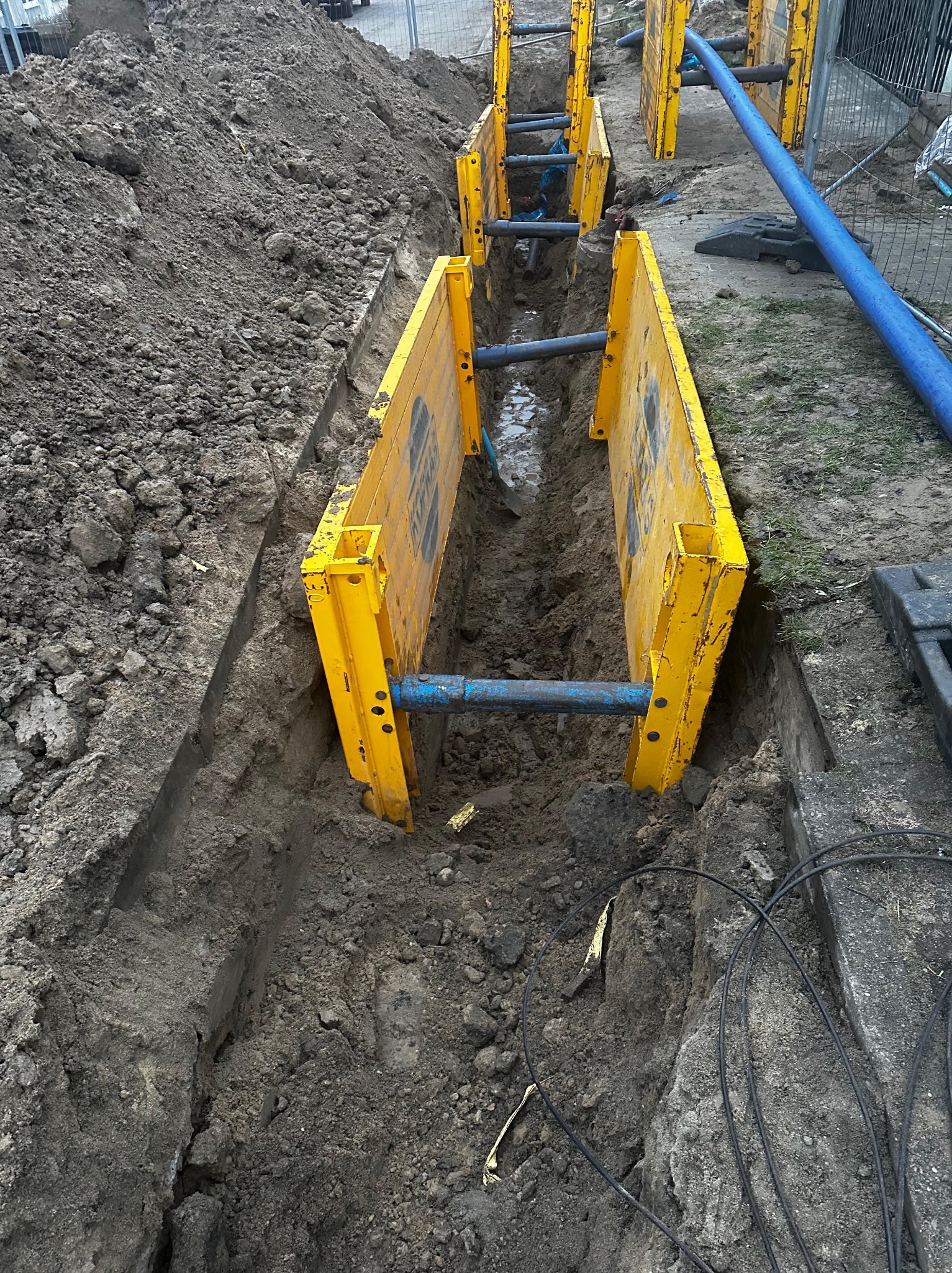 Wykop zabezpieczony żółtymi szalunkami stalowymi, widoczne niebieskie rury i hałda ziemi obok wykopu.