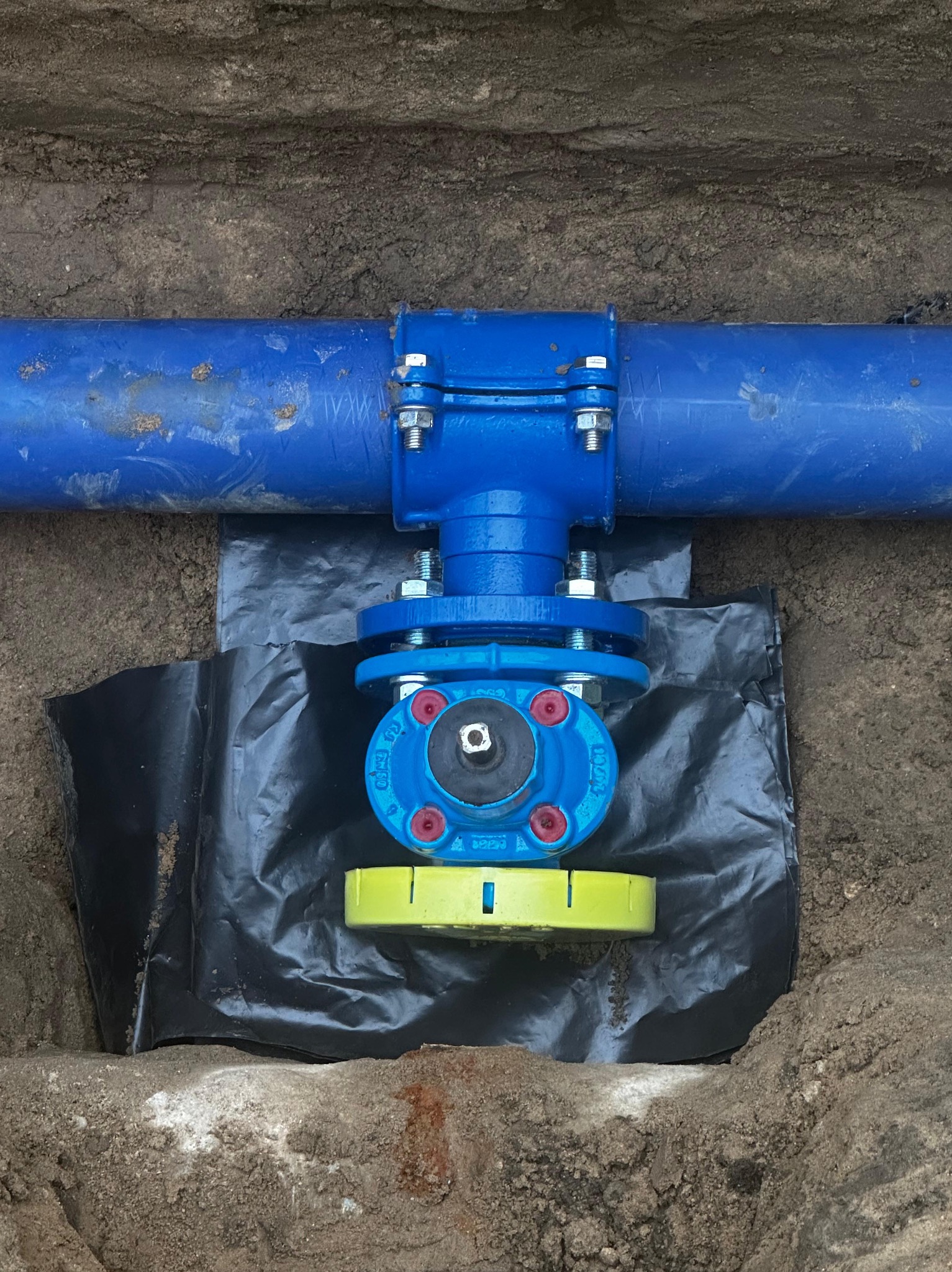 Niebieska rura wodociągowa z zaworem, zamontowana w wykopie, zabezpieczona czarną folią i poziomowana żółtą poziomicą.