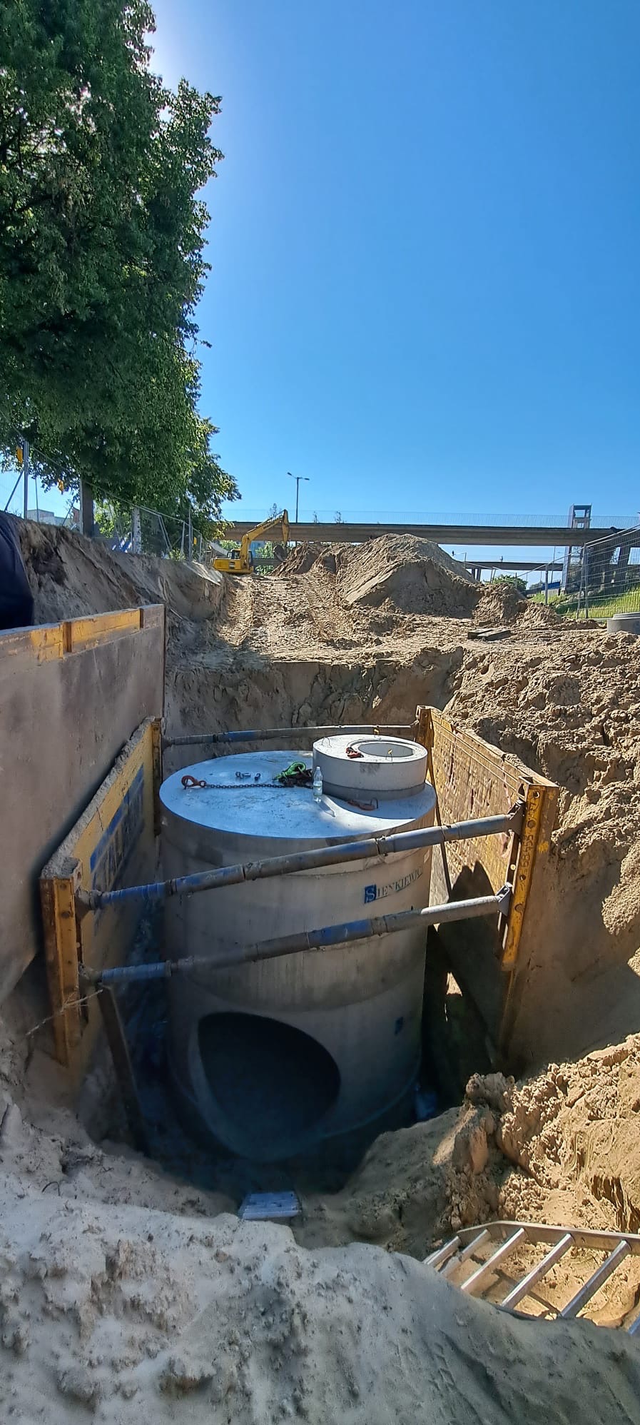 Betonowy zbiornik z otworem bocznym, zabezpieczony żółtymi szalunkami, umieszczony w wykopie ziemnym, z koparką widoczną w tle.