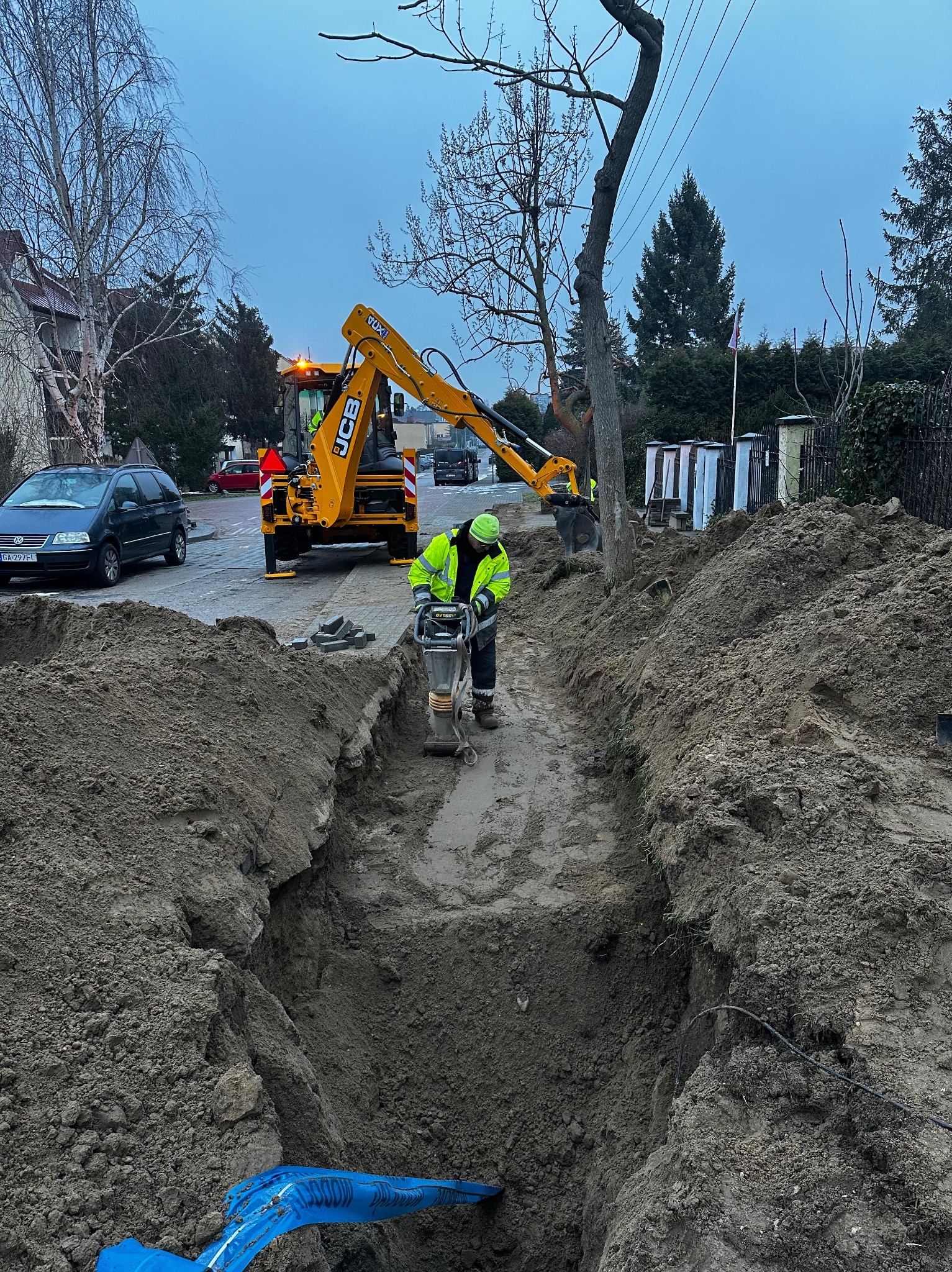 Pracownik w zielonej kamizelce zagęszcza ziemię w wykopie przy użyciu ubijarki spalinowej, w tle koparka JCB i ulica z samochodami.