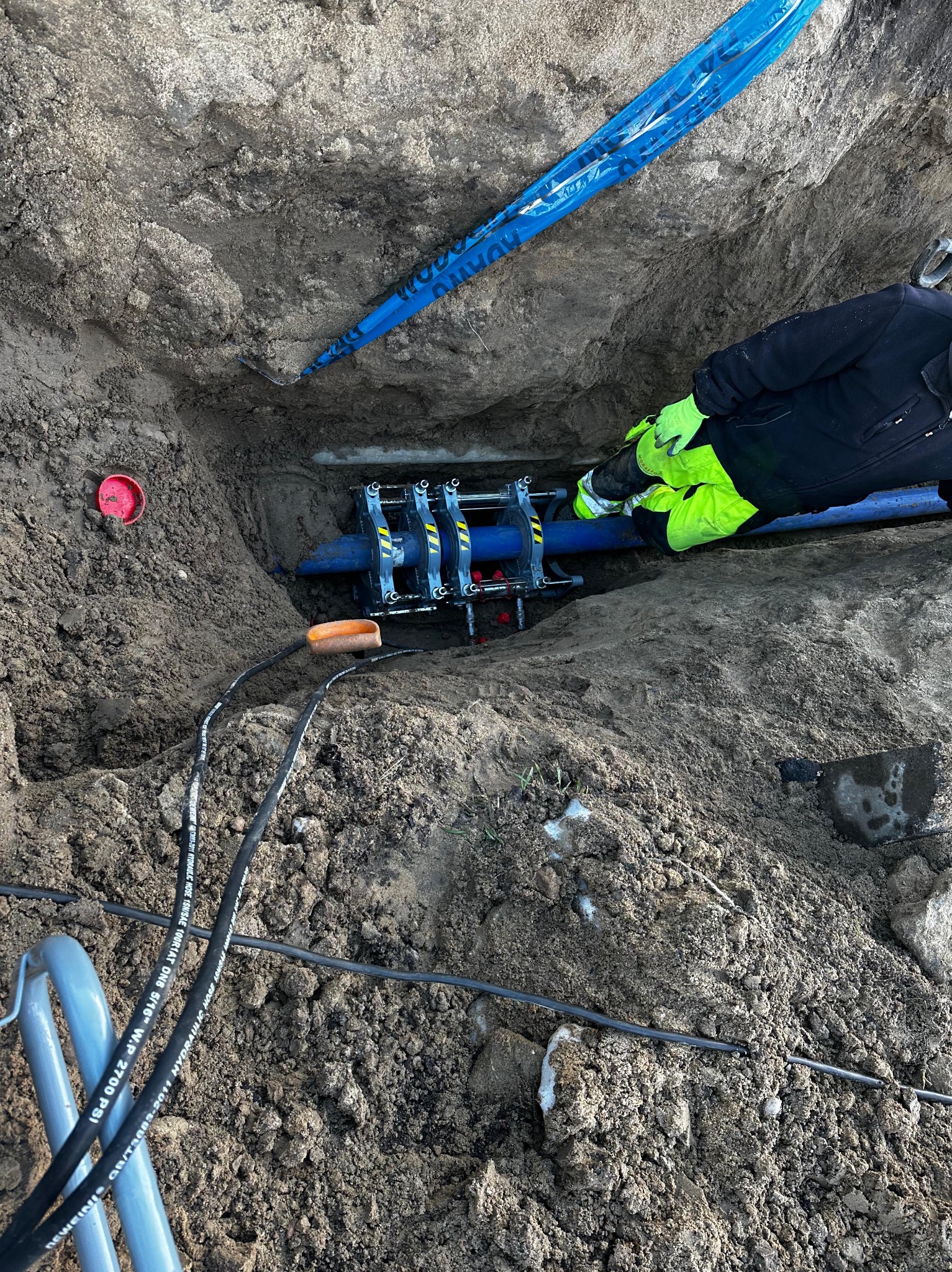 Pracownik w odblaskowej kurtce łączy rury wodociągowe w wykopie za pomocą specjalistycznego zaciskarki hydraulicznej, widoczne kable hydrauliczne i taśma ostrzegawcza.