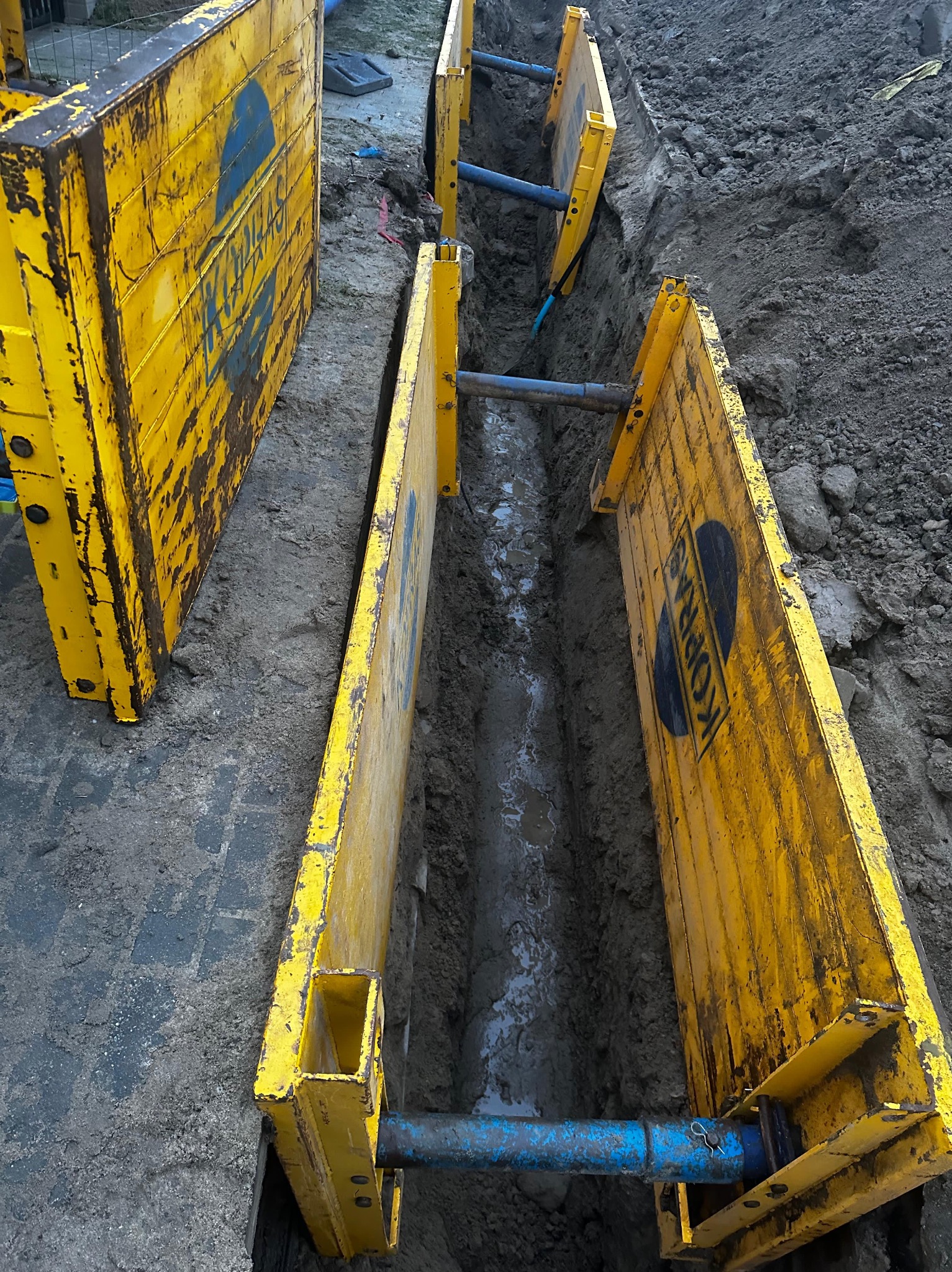 Głęboki wykop zabezpieczony żółtymi szalunkami stalowymi z widocznym niebieskim rurociągiem w ziemi, na krawędzi wykopu widoczna kostka brukowa.