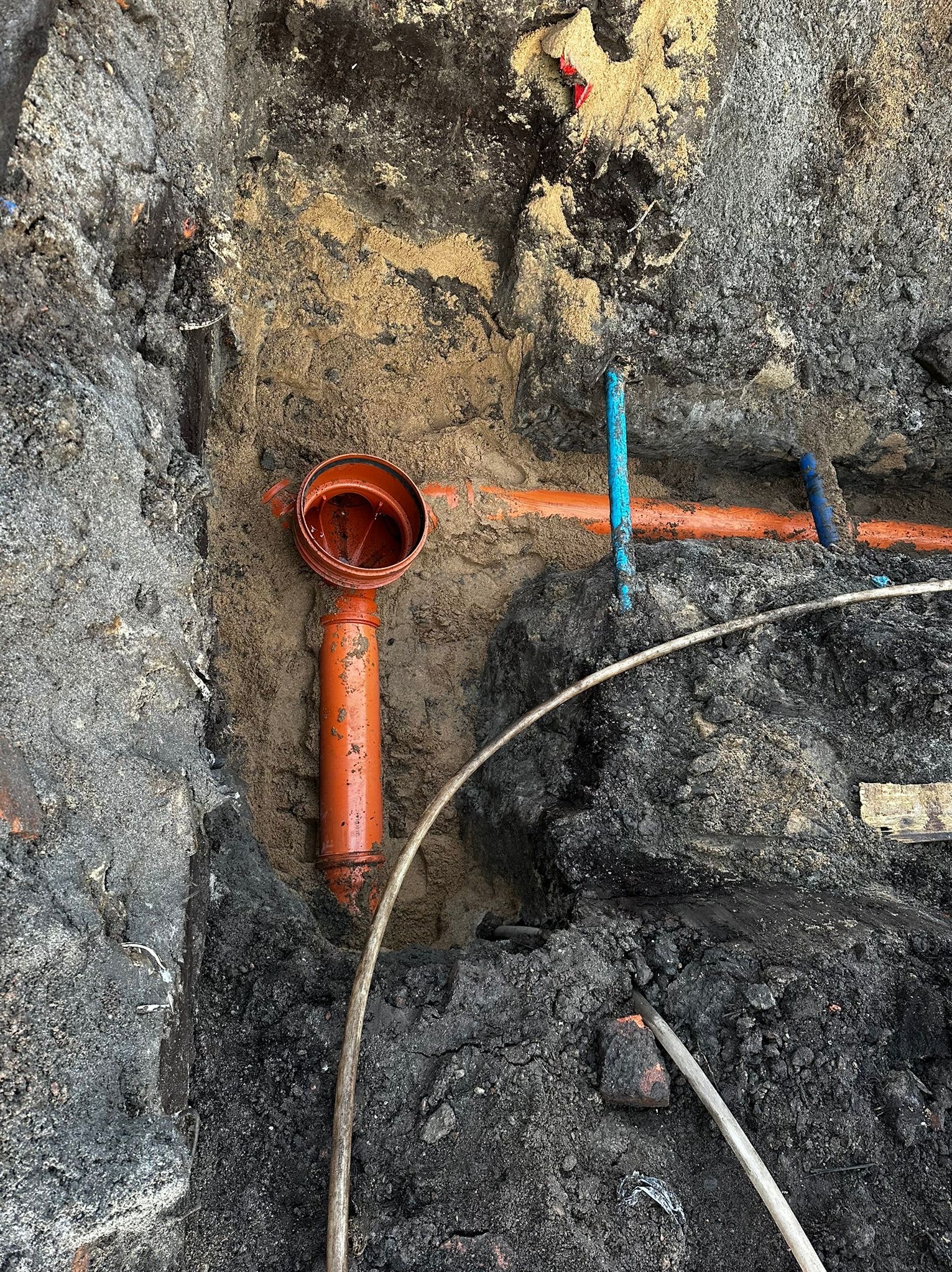 Wykop z widocznymi rurami kanalizacyjnymi z tworzywa sztucznego, pomarańczowa pionowa rura z kielichem skierowanym do góry, niebieskie rury poziome.