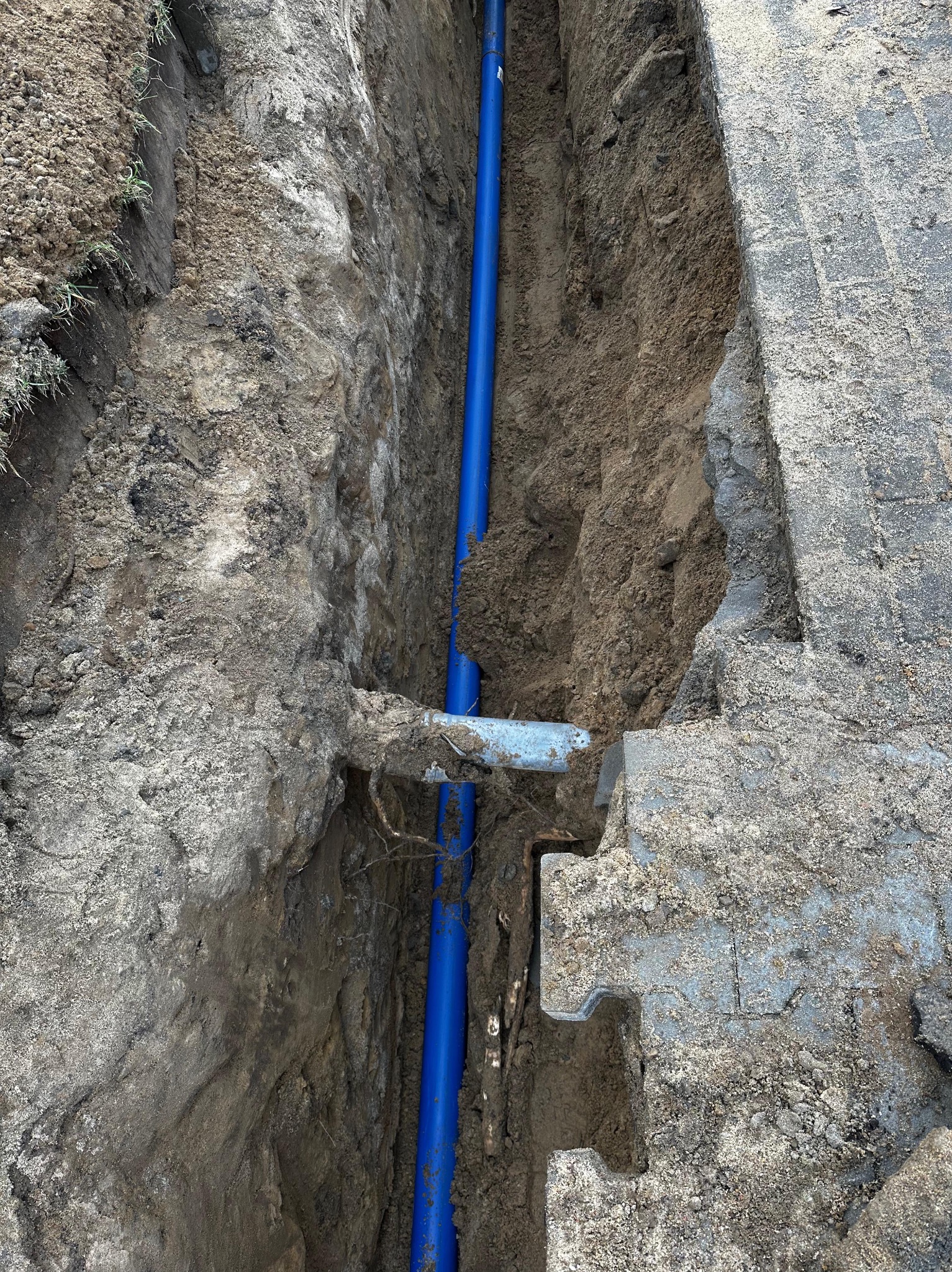 Wykop z ułożonym niebieskim rurociągiem wodnym, widoczne przyłącze i fragment betonowej konstrukcji z wycięciami, ziemia gliniasta.