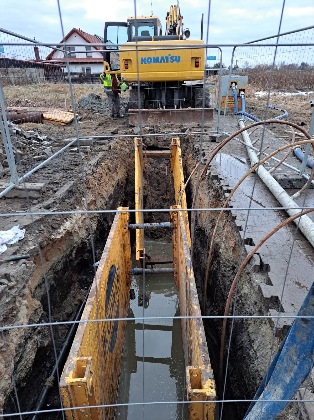 Głęboki wykop zabezpieczony żółtymi szalunkami, wypełniony wodą, z pracującą koparką Komatsu w tle i widocznym pracownikiem w odblaskowej kamizelce, teren budowy ogrodzony siatką.