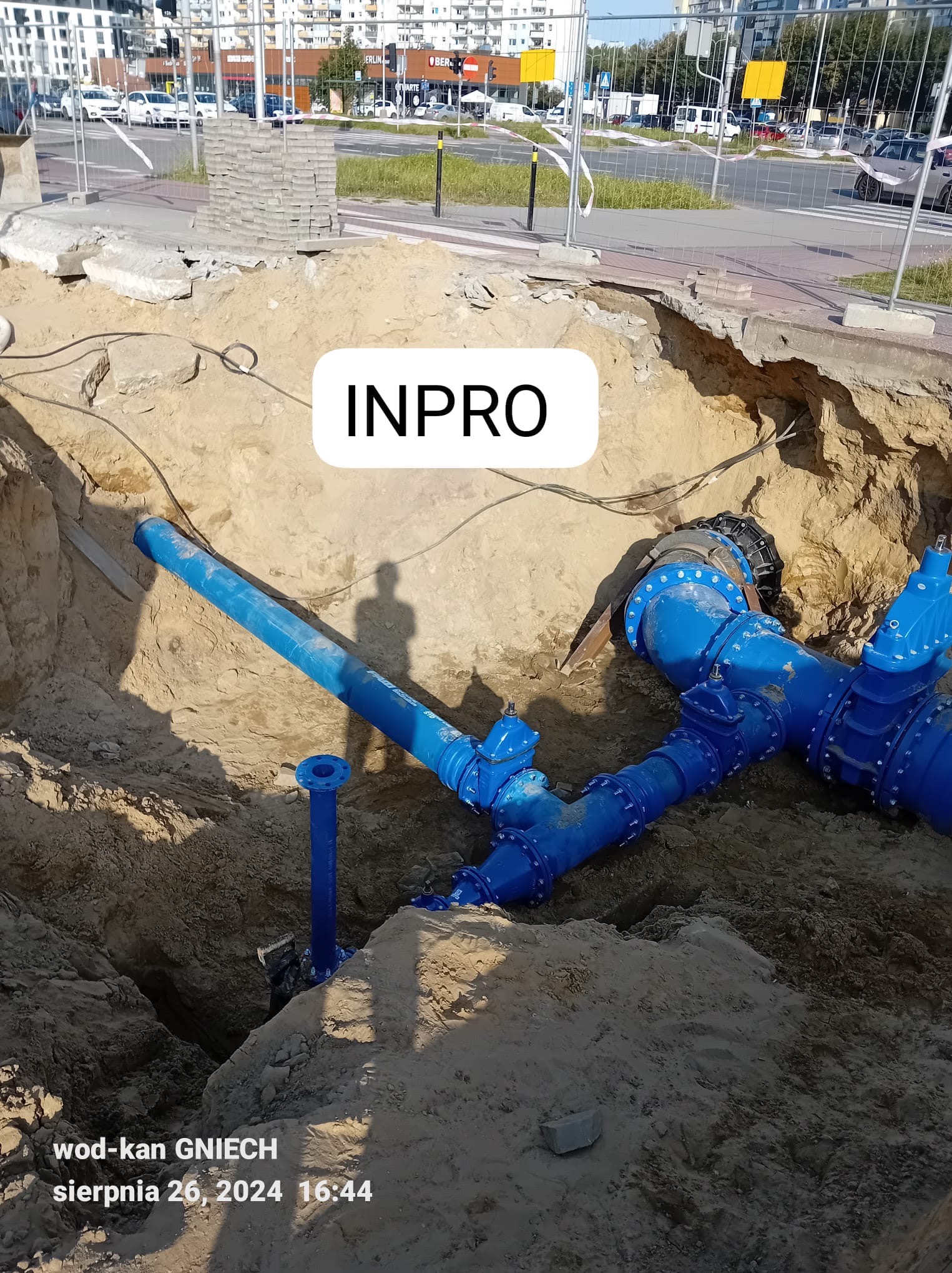 Wykop z widocznym, częściowo zamontowanym systemem niebieskich rur wodociągowych z licznymi złączkami i zaworami, widoczne ślady ziemi, w tle ulica z chodnikiem i budynkami.