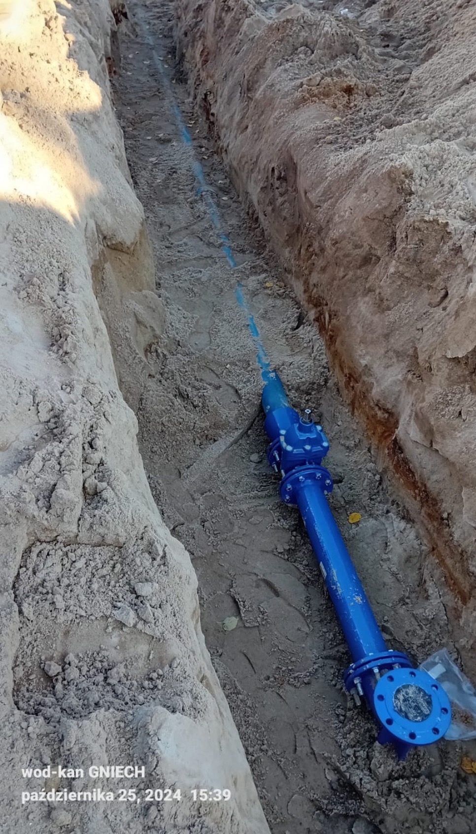 Niebieska rura wodociągowa z zaworem ułożona w wykopie ziemnym o piaszczystych ścianach, widoczna linia rury biegnąca wzdłuż wykopu.