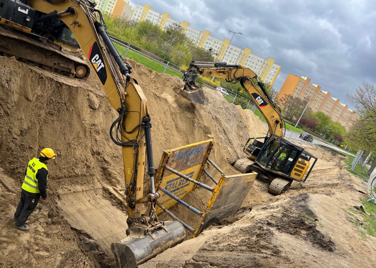 Dwa żółte koparki CAT pracują w wykopie w Pruszczu Gdańskim, widoczny pracownik w żółtym kasku i kamizelce odblaskowej oraz zabezpieczenia wykopu.