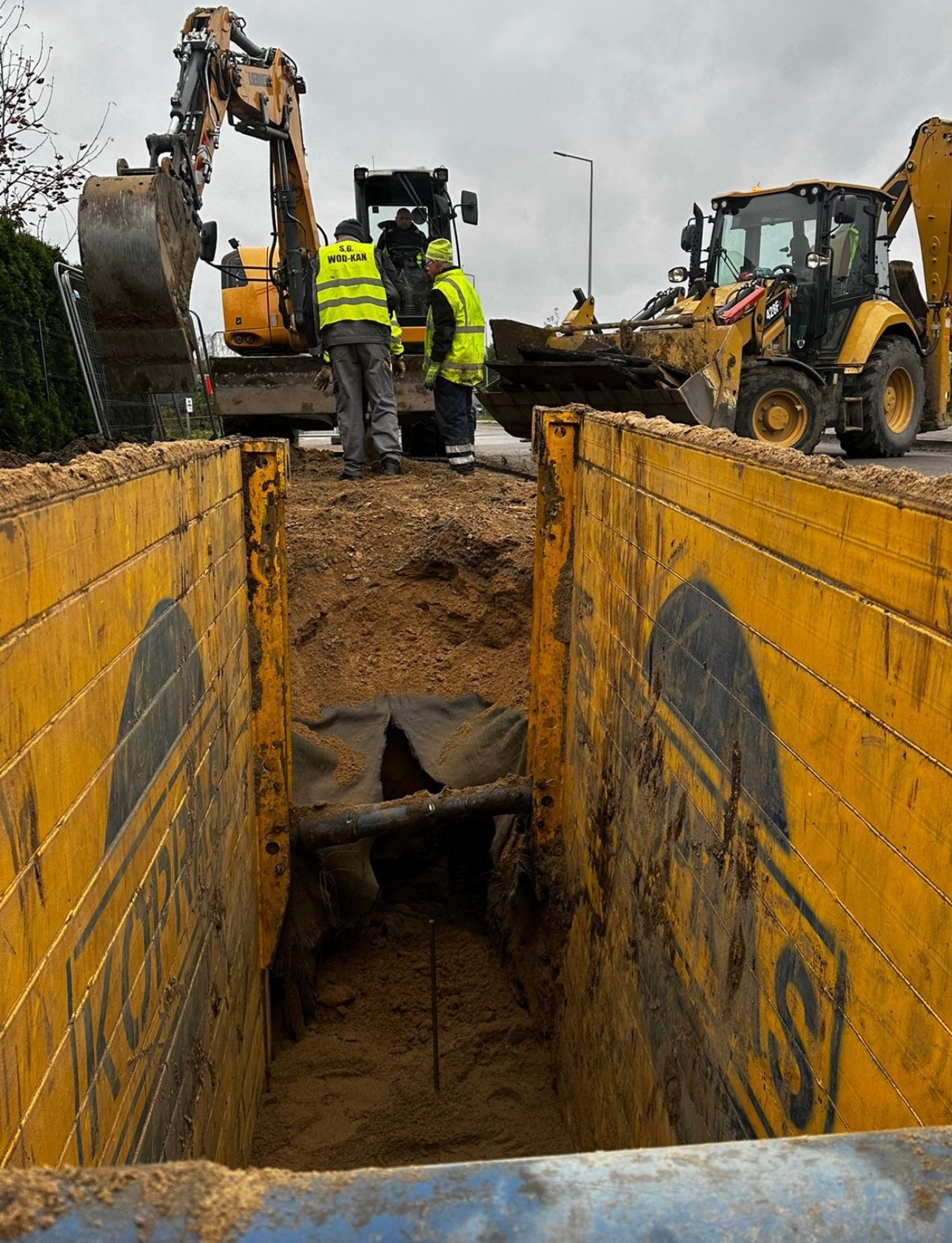 Wykop zabezpieczony żółtymi szalunkami, wewnątrz widoczna rura i fragment geowłókniny, na zewnątrz pracują dwie koparki i trzech pracowników w odblaskowych kamizelkach z logo firmy.