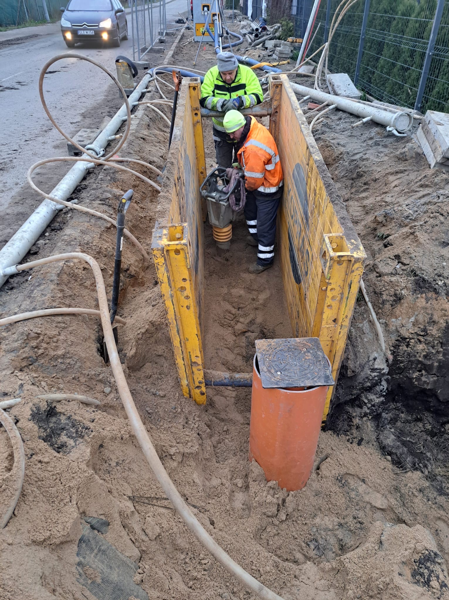 Prace ziemne przy wykopie zabezpieczonym żółtymi szalunkami, dwóch robotników w odblaskowych ubraniach, pomarańczowa studzienka kanalizacyjna z żeliwnym deklem, widoczne rury i węże na powierzchni...