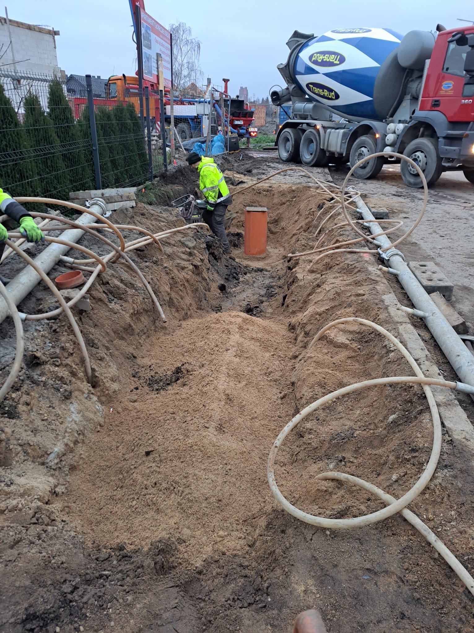 Wykop z rurami i pomarańczową studzienką podczas instalacji przyłącza wodno-kanalizacyjnego, w tle betoniarka Trans-Rud i maszyny budowlane.