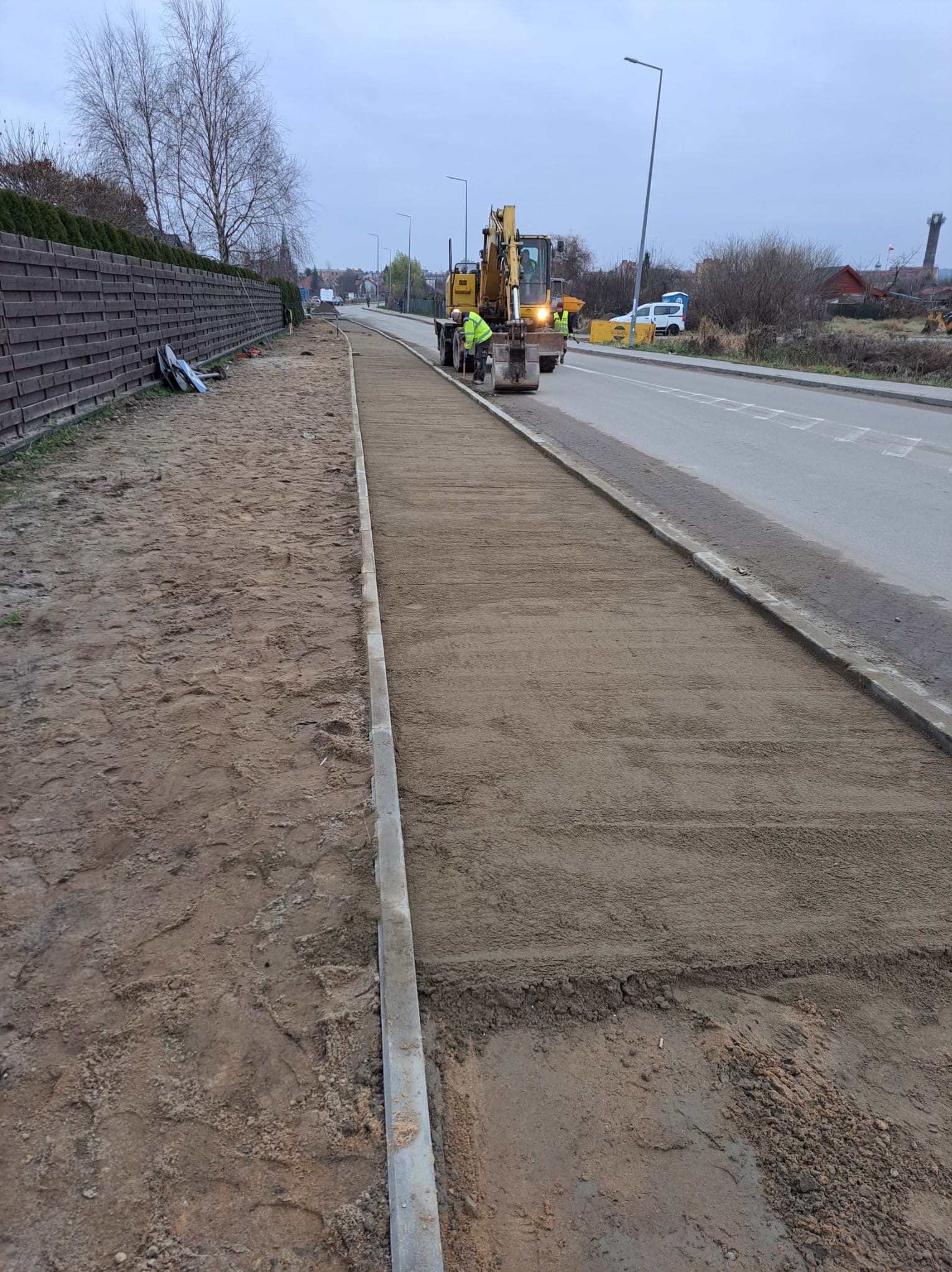 Przygotowanie podłoża pod chodnik z kostki brukowej, wyrównany piasek i obrzeża z kostki betonowej, w tle koparka i pracownicy.