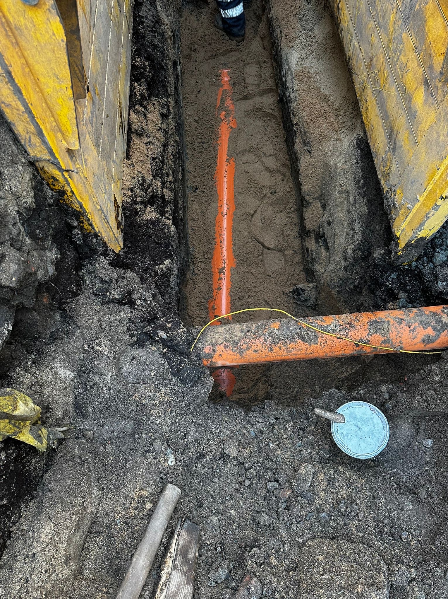 Wykonany wykop zabezpieczony żółtymi szalunkami, w którym ułożono pomarańczowe rury, widoczne narzędzia i ziemia.