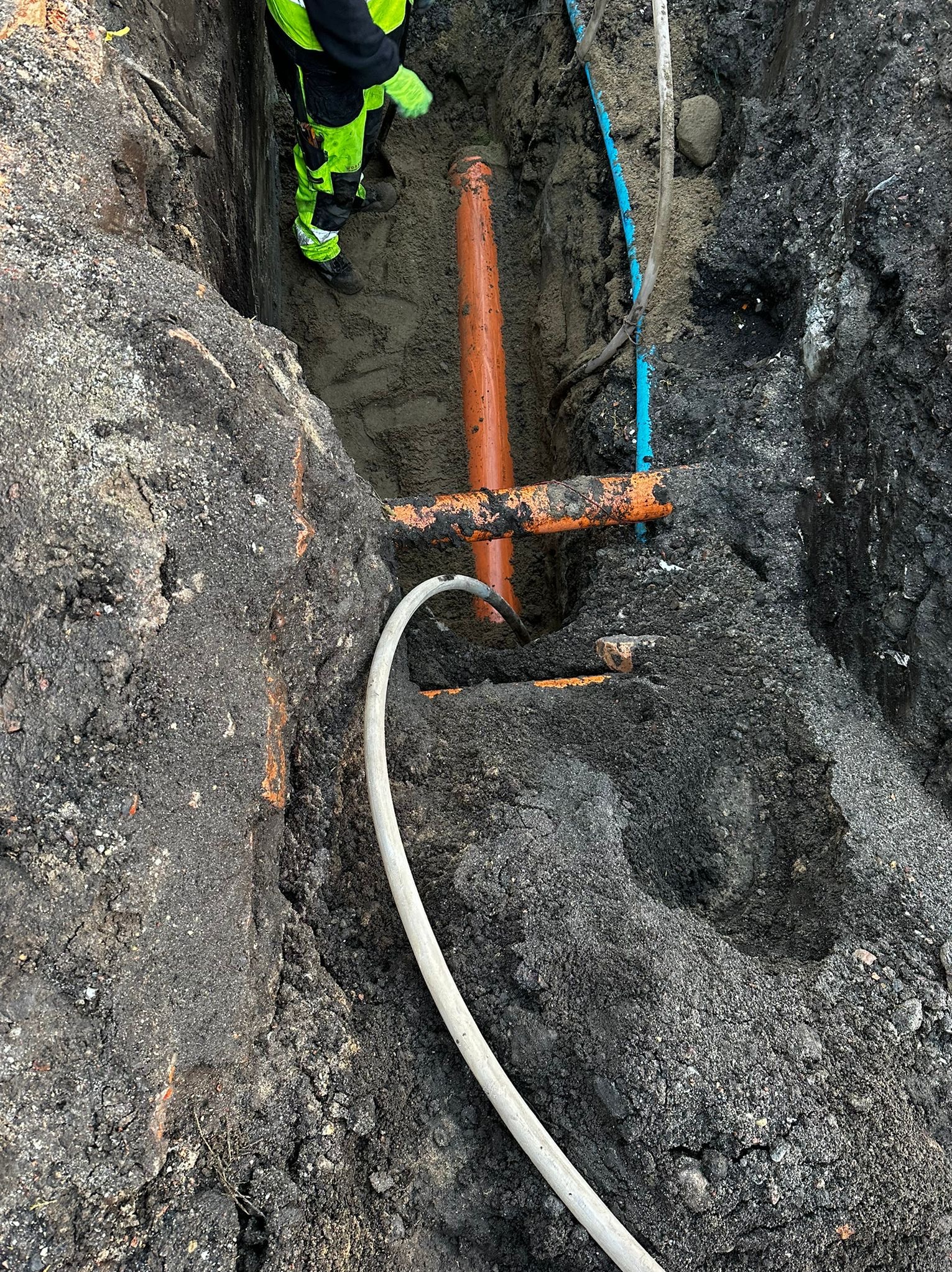 Głęboki wykop z widocznymi pomarańczowymi i niebieskimi rurami instalacyjnymi oraz fragmentem węża, pracownik w odblaskowym ubraniu w tle.