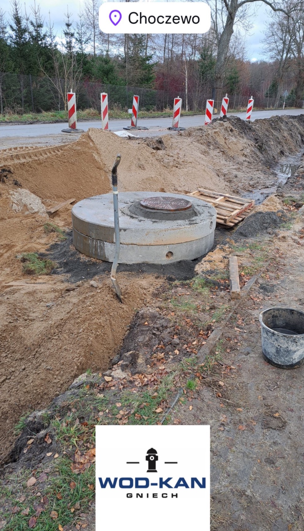 Wkopany betonowy krąg z włazem kanalizacyjnym, obok łopata wbita w ziemię, widoczne zabezpieczenia drogowe i logo firmy WOD-KAN Gniech.