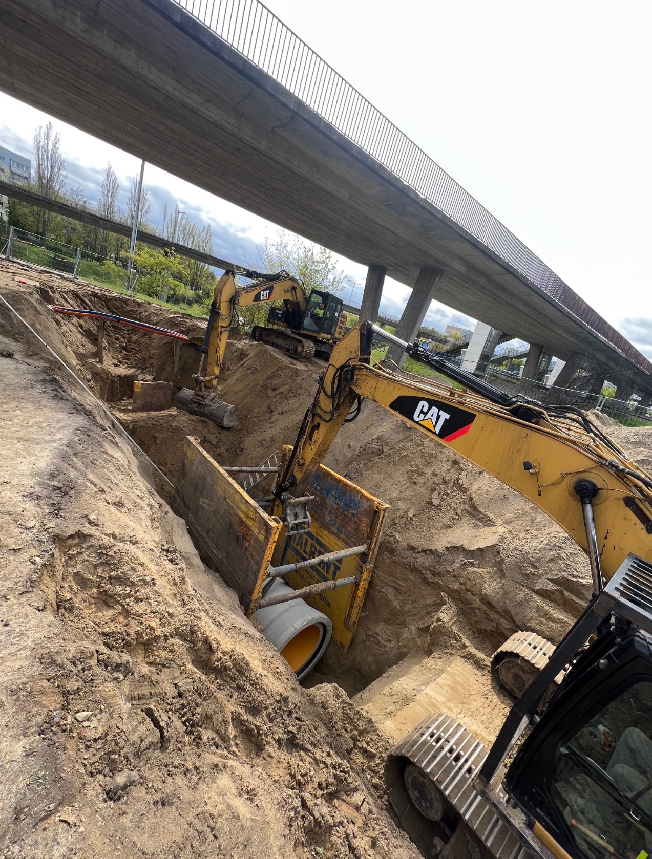 Głęboki wykop pod estakadą, zabezpieczony żółtymi szalunkami, z widocznymi rurami i dwoma koparkami CAT, sugerujący prace ziemne związane z instalacją podziemną.