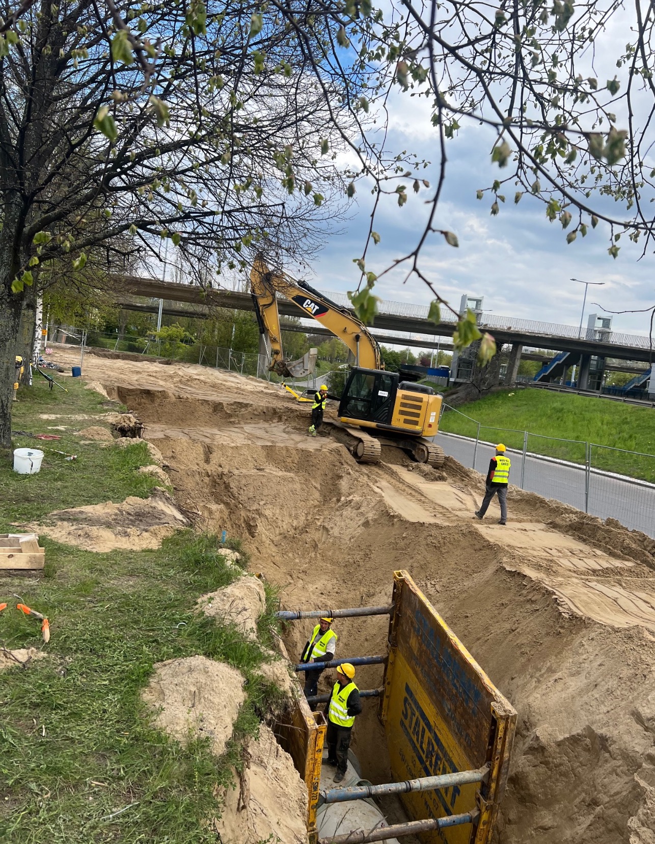 Wykop zabezpieczony żółtymi szalunkami, wewnątrz pracownicy w kaskach i kamizelkach odblaskowych, na górze stoi koparka CAT i kolejny pracownik, w tle widoczny wiadukt.