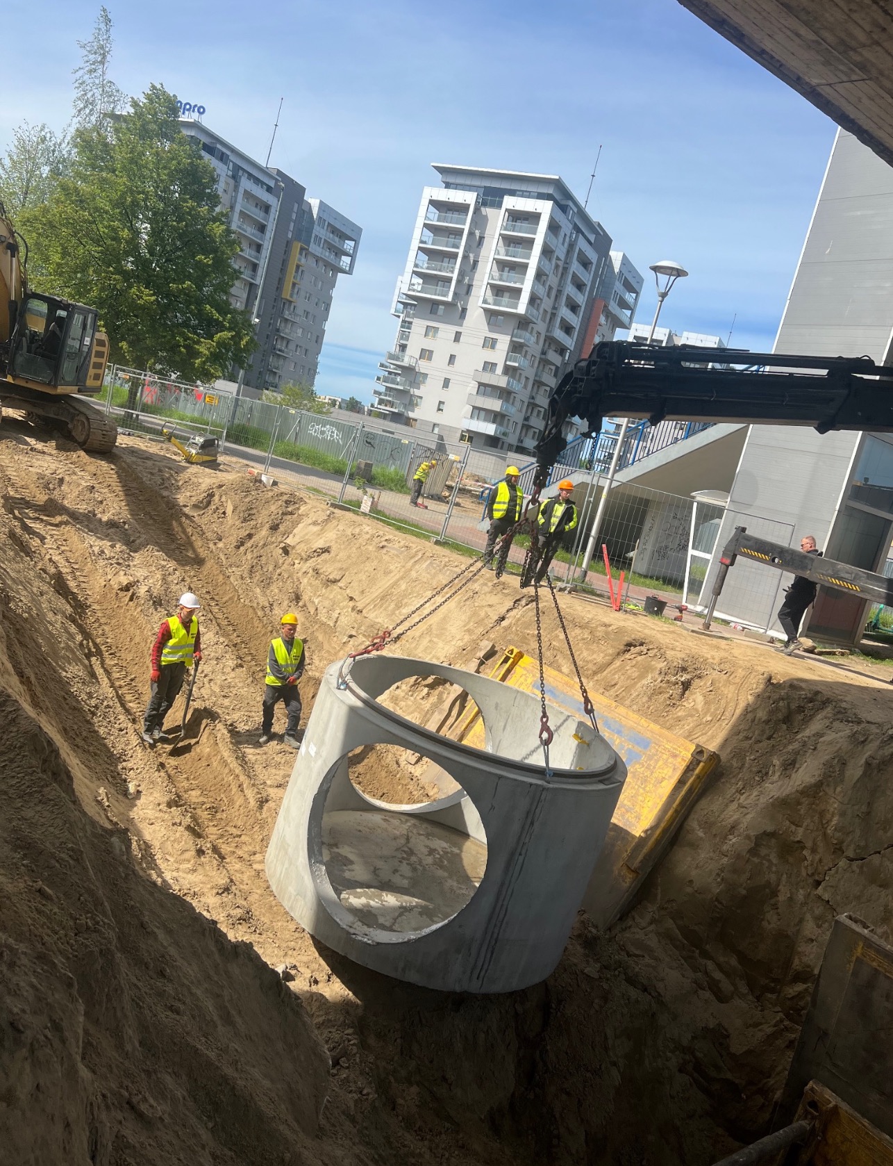 Montaż betonowego kręgu z otworami na rury w wykopie, z użyciem dźwigu, pracownicy w kamizelkach odblaskowych, na tle budynki mieszkalne.
