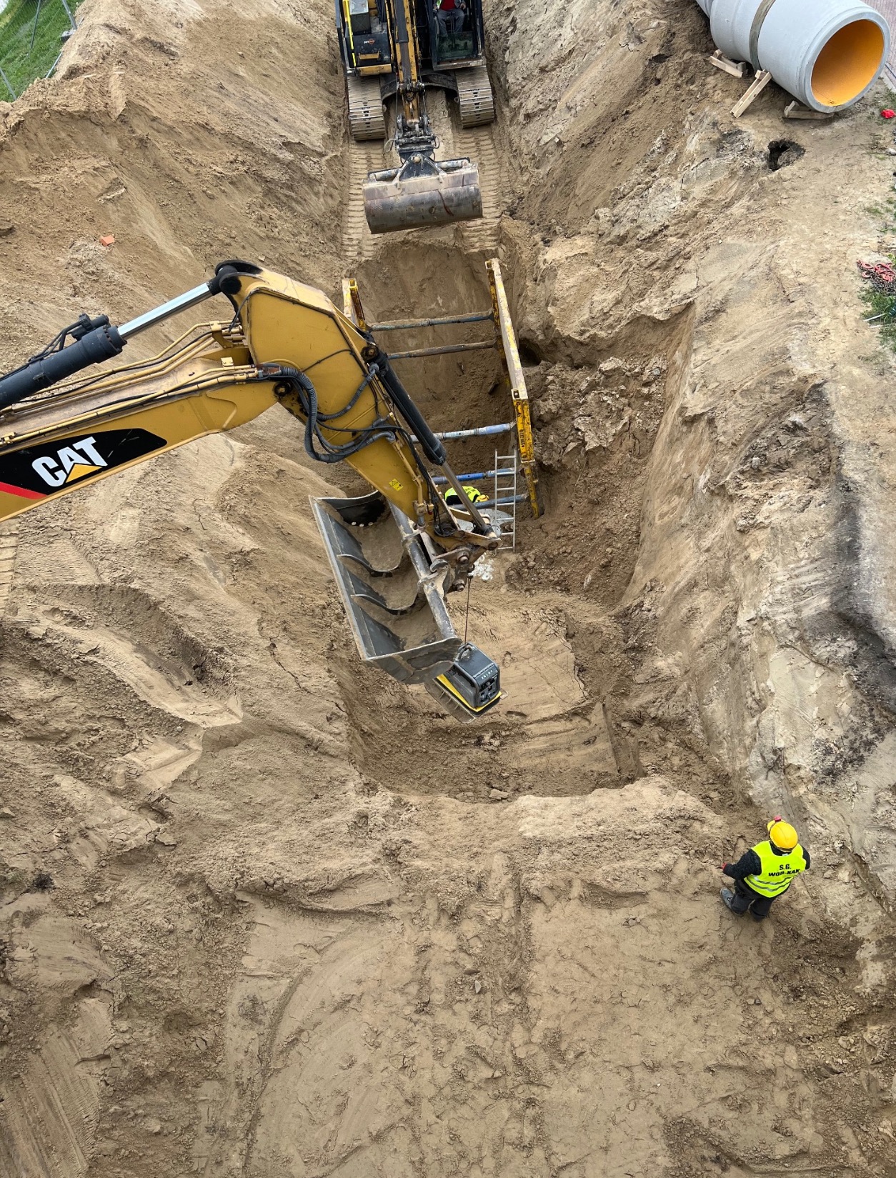 Głęboki wykop z zabezpieczeniami ścian, dwie koparki CAT pracują wewnątrz, a pracownik w żółtej kamizelce mierzy teren w rogu wykopu. W tle widoczne rury.