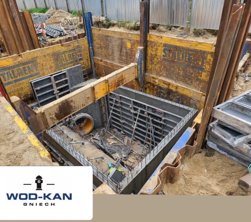 Wykop zabezpieczony stalowymi szalunkami, widoczne rury kanalizacyjne i elementy konstrukcyjne w trakcie montażu.