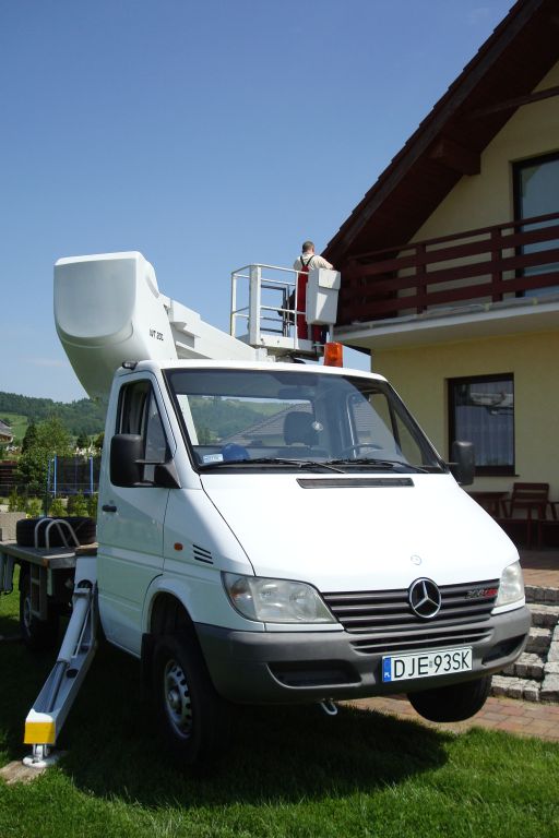 Biały podnośnik koszowy Mercedes Sprinter z wysięgnikiem podniesionym do dachu jednopiętrowego domu; pracownik w czerwonym kombinezonie na platformie roboczej wykonuje prace na wysokości.
