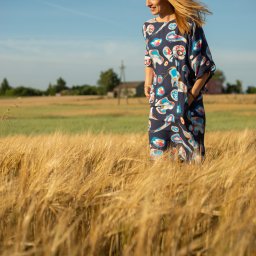 Visual Works Studio - Portret kobiety w kwiecistej sukience na tle pola zboża i błękitnego nieba. Uśmiechnięta, z rozwianymi włosami, patrzy w bok. Naturalne światło, ciepłe kolory.