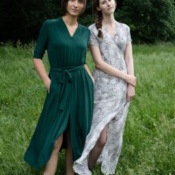 Visual Works Studio - Dwie kobiety w sukienkach na łące: brunetka w szmaragdowej sukni z rozcięciem i blondynka w sukience w cętki. Sesja w plenerze, naturalne światło, swobodna atmosfera.
