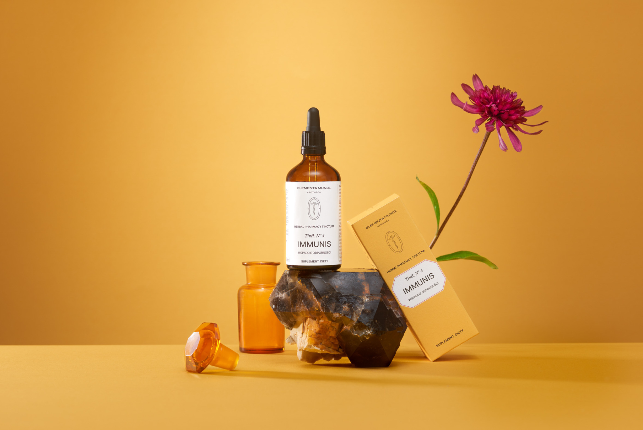 Zdjęcie produktowe: butelka z pipetą, pudełko suplementu Elementa Mundi, flakonik i kwiat na abstrakcyjnym, pomarańczowym tle. Minimalistyczna kompozycja.