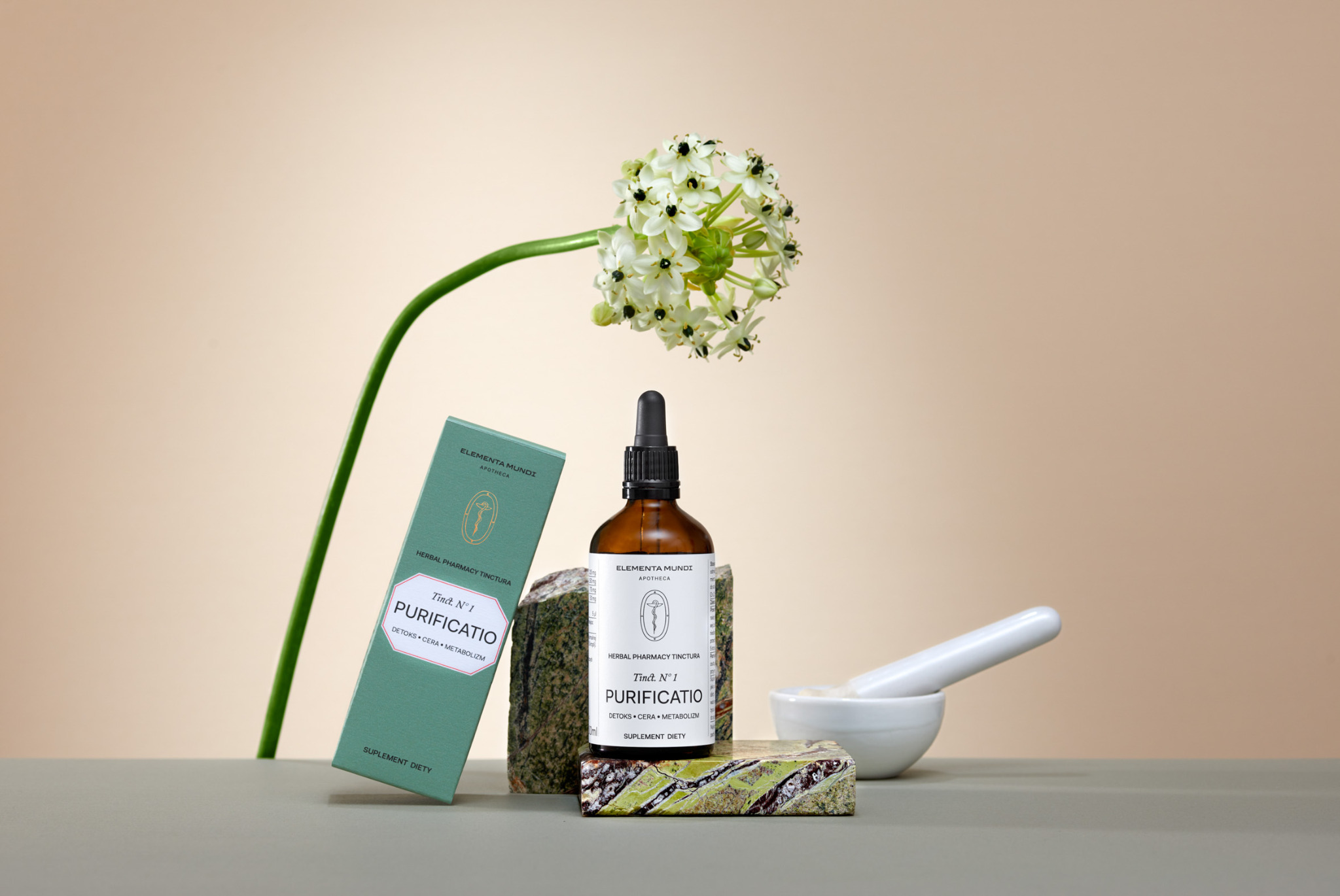 Zdjęcie produktowe Elementa Mundi: butelka z pipetą, pudełko i moździerz z tłuczkiem na tle beżowym, z kwiatem. Naturalne światło, minimalistyczna kompozycja.