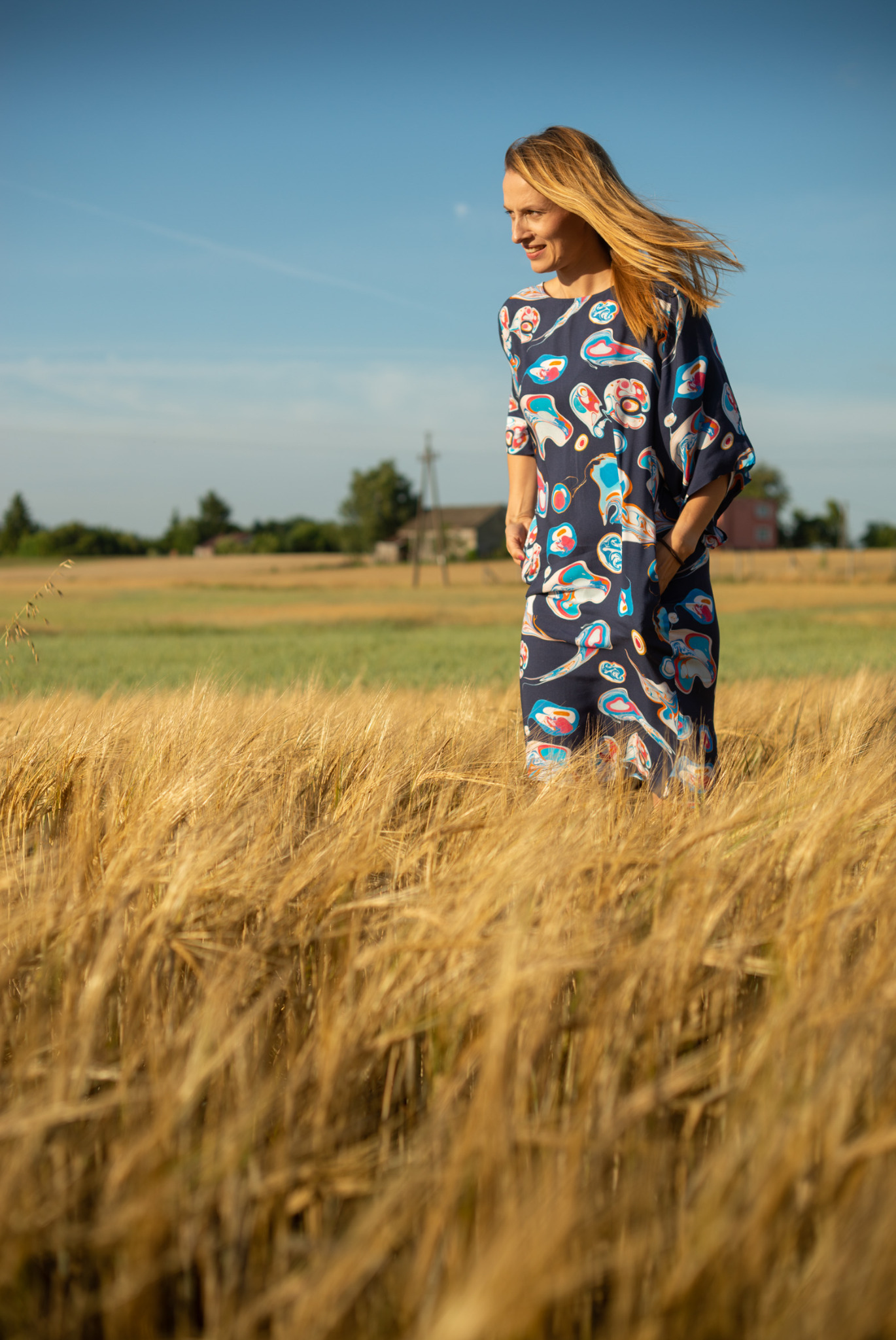 Portret kobiety w kwiecistej sukience na tle pola zboża i błękitnego nieba. Uśmiechnięta, z rozwianymi włosami, patrzy w bok. Naturalne światło, ciepłe kolory.