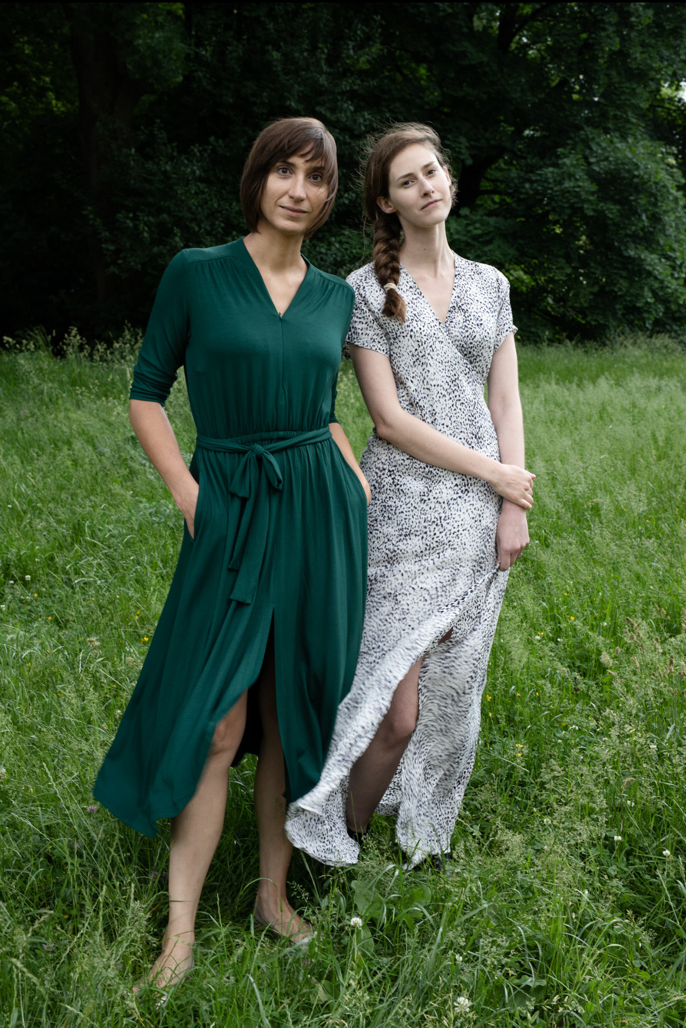 Dwie kobiety w sukienkach na łące: brunetka w szmaragdowej sukni z rozcięciem i blondynka w sukience w cętki. Sesja w plenerze, naturalne światło, swobodna atmosfera.