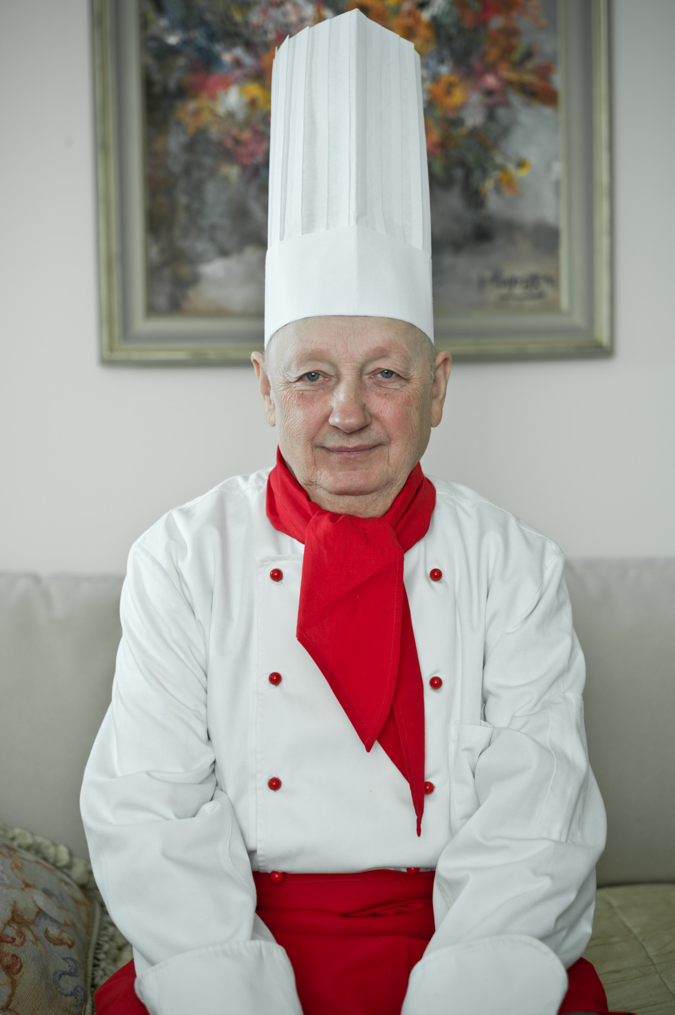 Portret uśmiechniętego starszego mężczyzny w stroju kucharza na tle jasnej ściany z obrazem. Czerwony krawat i guziki dodają koloru. Ujęcie centralne, dobre oświetlenie.