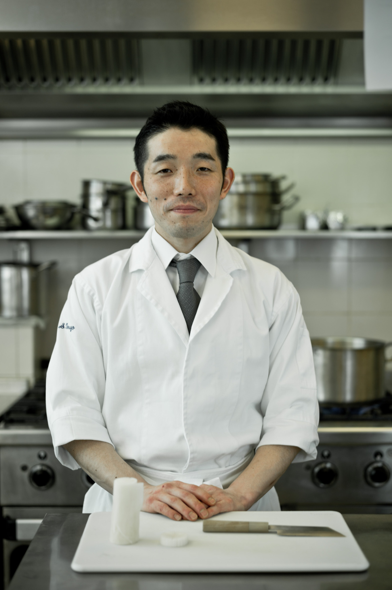 Portret kucharza w białym uniformie na tle profesjonalnej kuchni, z deską i nożem na pierwszym planie. Uśmiechnięty, z rękami złożonymi na desce.