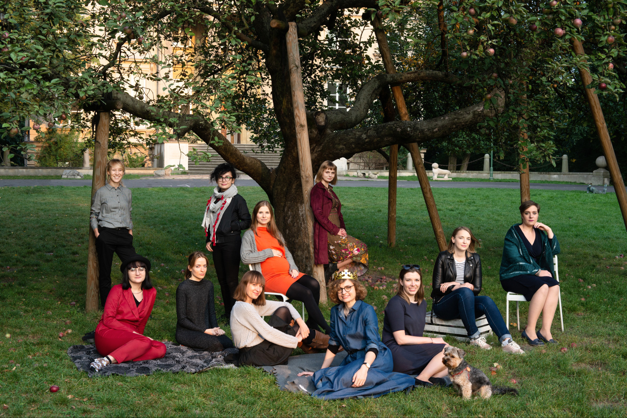 Grupowy portret plenerowy jedenastu kobiet i psa pod jabłonią wspartą drewnianymi belkami, w tle budynek z kolumnami i rzeźby lwów.